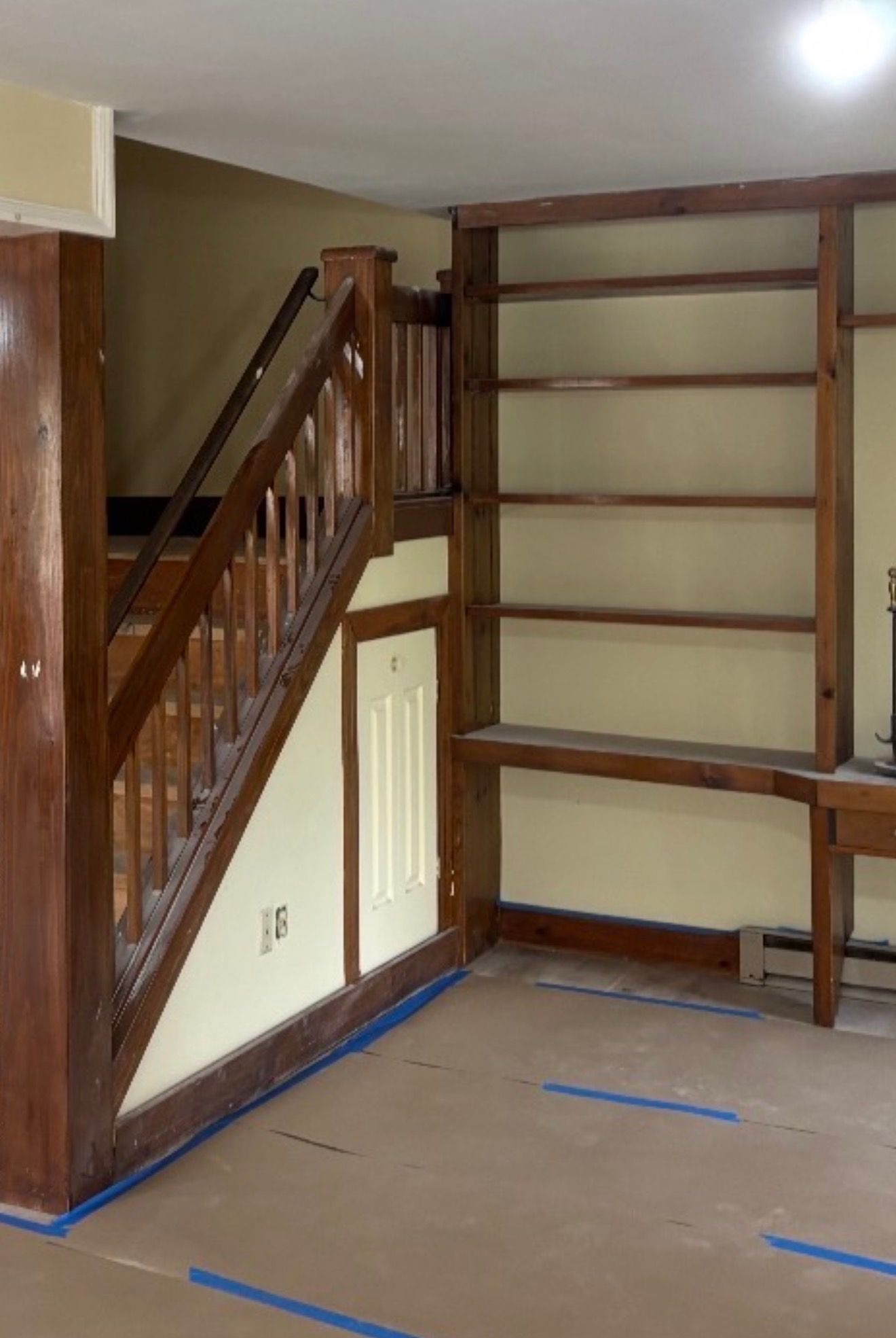 A wooden staircase with a built-in door and shelving unit in a room with protective paper flooring and blue painter's tape.