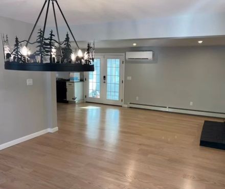 A spacious room with light hardwood floors, a decorative forest-themed chandelier, French doors, and a wall-mounted AC unit.