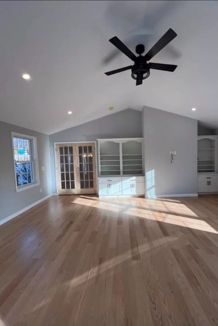A sunlit living room with vaulted ceilings, light wood floors, gray walls, a ceiling fan, and white built-in shelving.