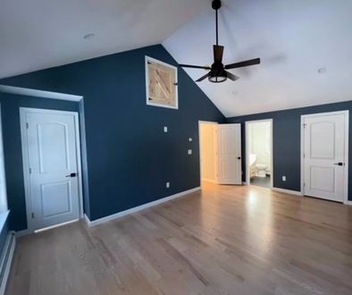 A bright, empty room with dark blue walls, light wood flooring, three white doors, and a ceiling fan.