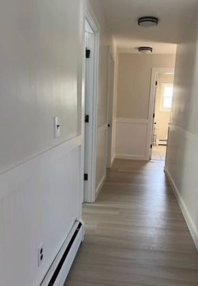A hallway with light-colored flooring, beige walls with white wainscoting, and recessed lighting leading to a doorway.