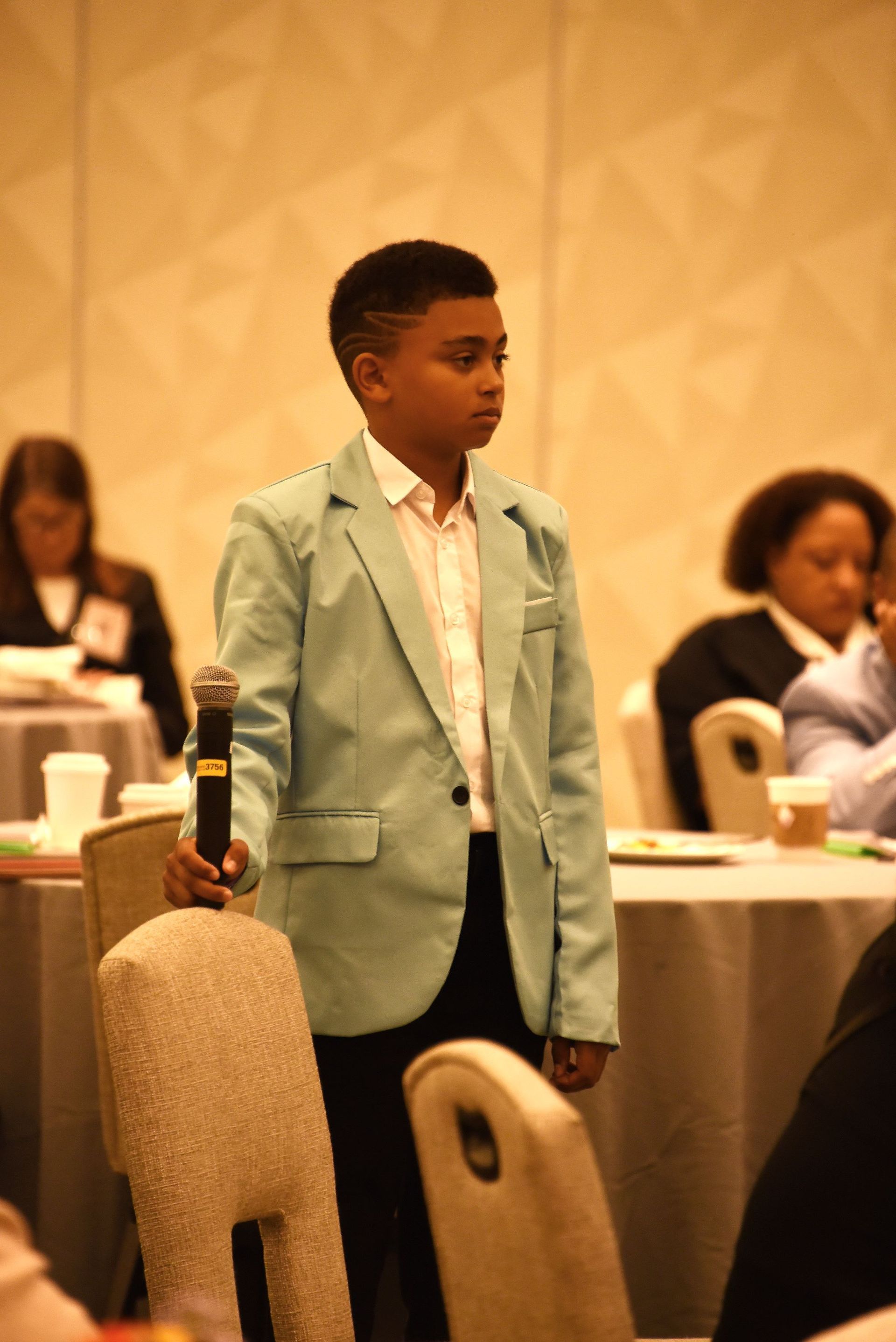 A young boy in a green jacket is holding a microphone in front of a group of people.