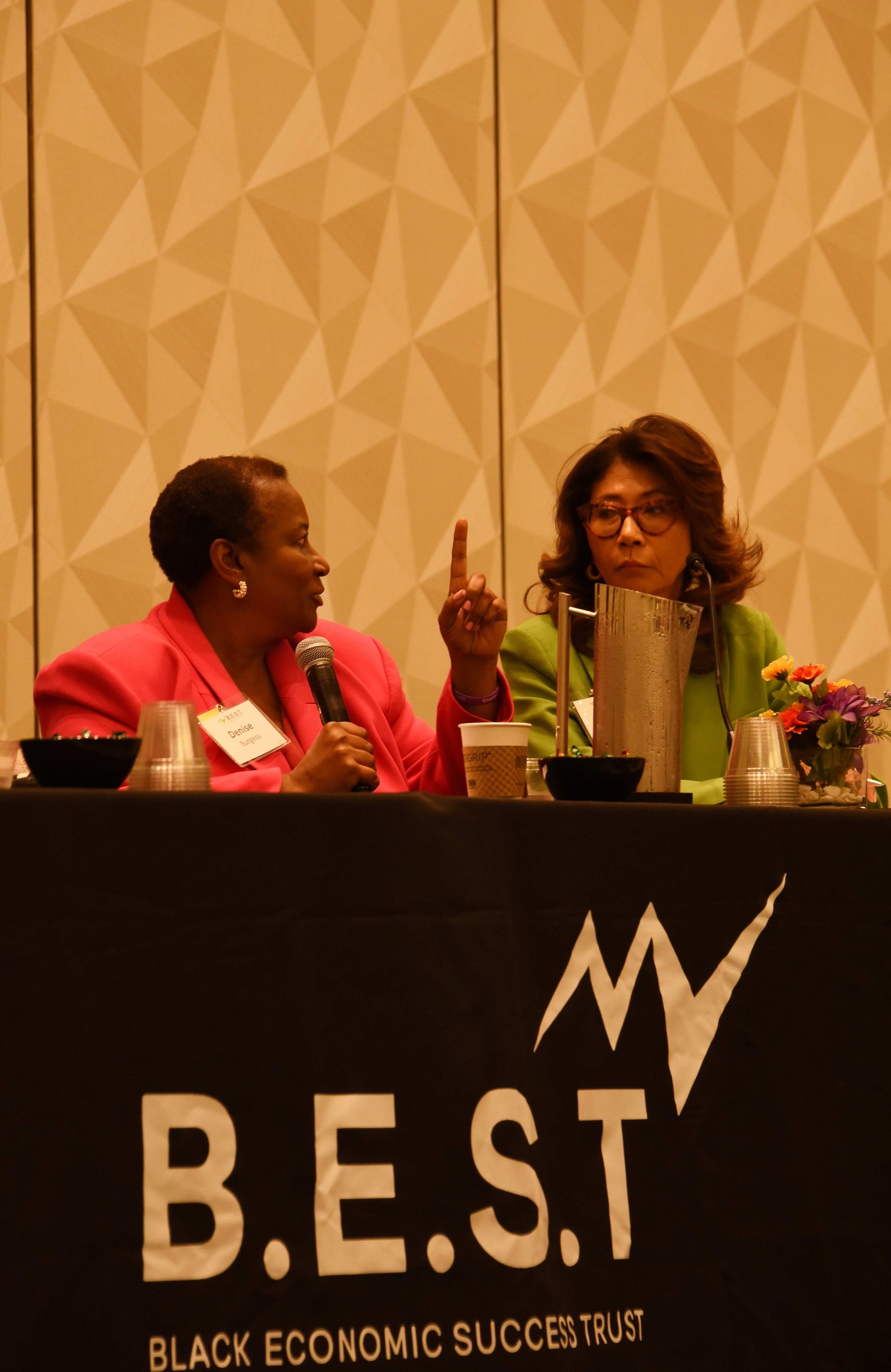 Two women sit at a table with a sign that says b.e.s.t.