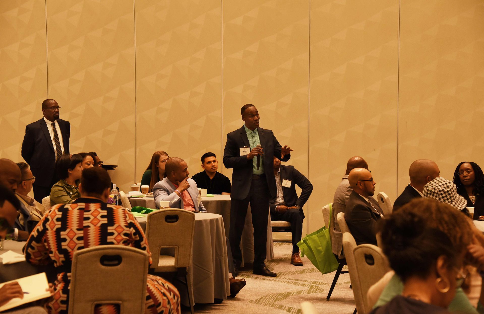 A man in a suit is standing in front of a group of people sitting at tables.