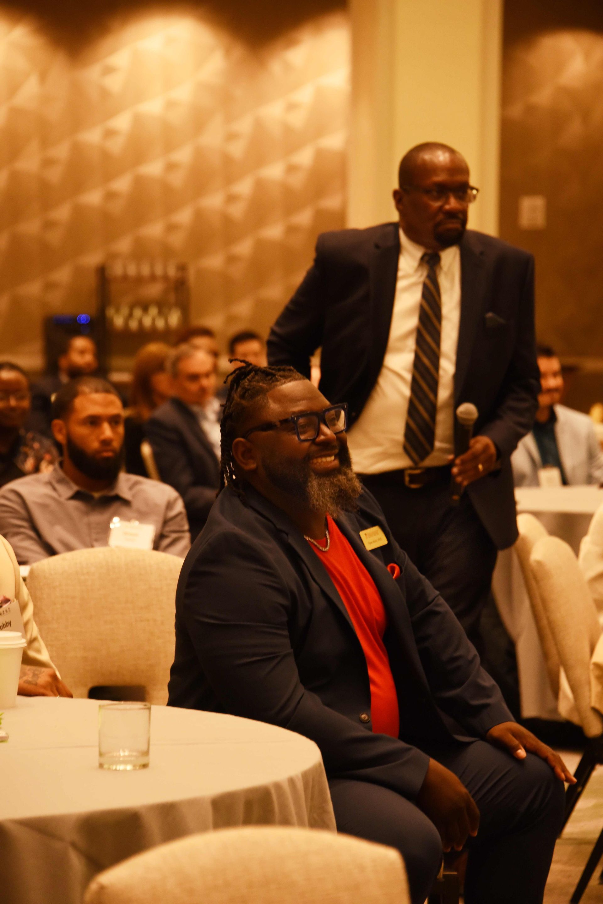 A man in a suit and tie is standing in front of a group of people sitting at tables.