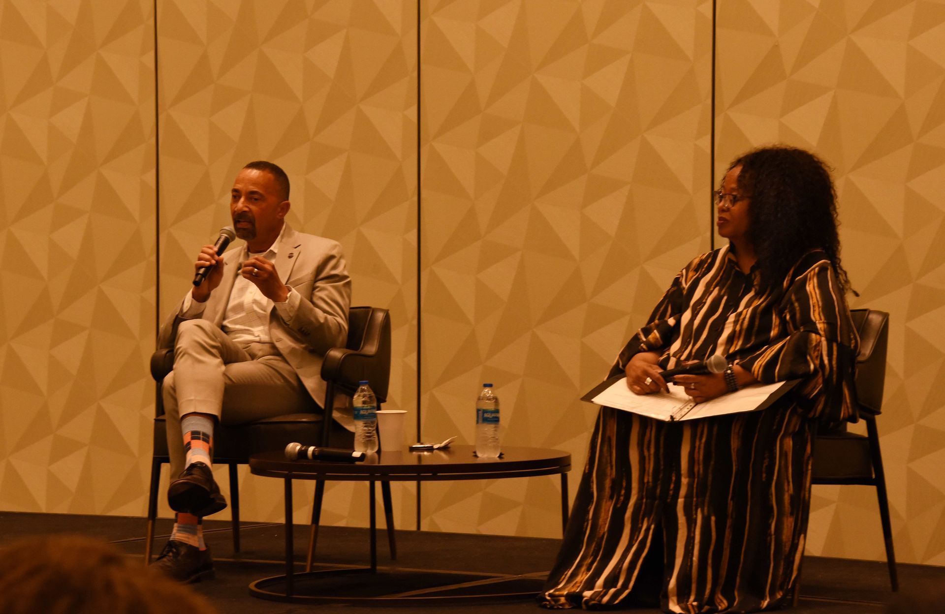 A man is sitting in a chair talking into a microphone while a woman sits at a table.