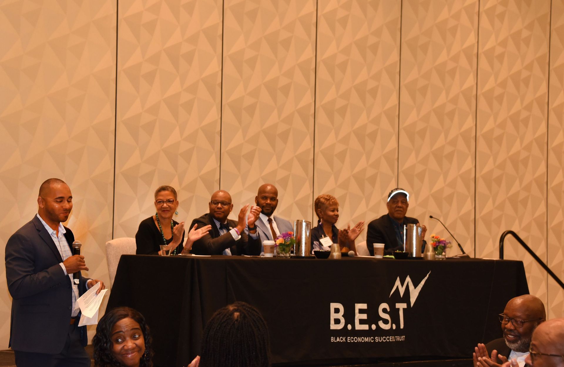A group of people are sitting at a table in front of a b.e.s.t. sign.