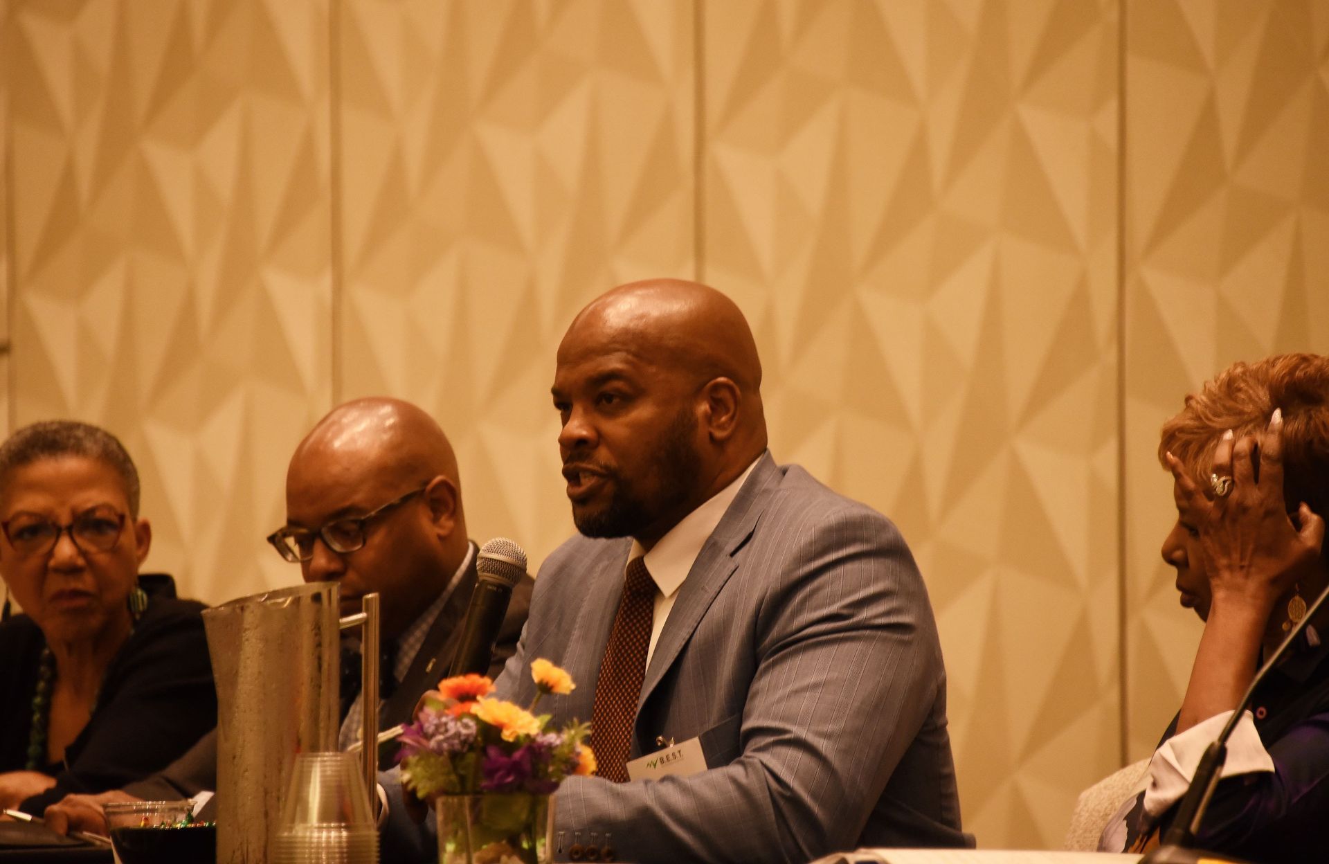 A man in a suit and tie is sitting at a table talking to a group of people.