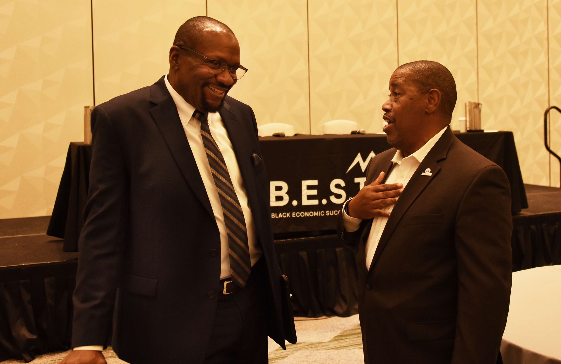 Two men in suits are talking in front of a sign that says b.e.s.t.