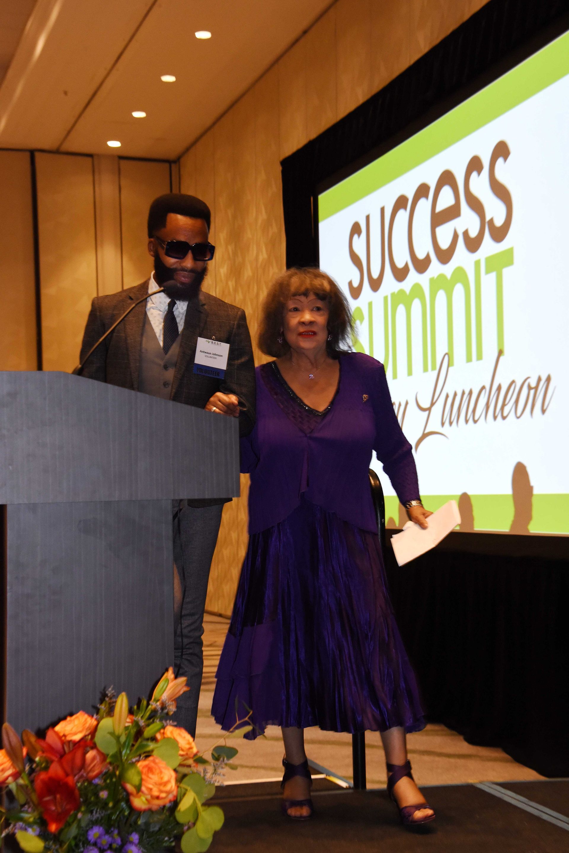 A woman in a purple dress stands next to a man at a podium at a success summit luncheon