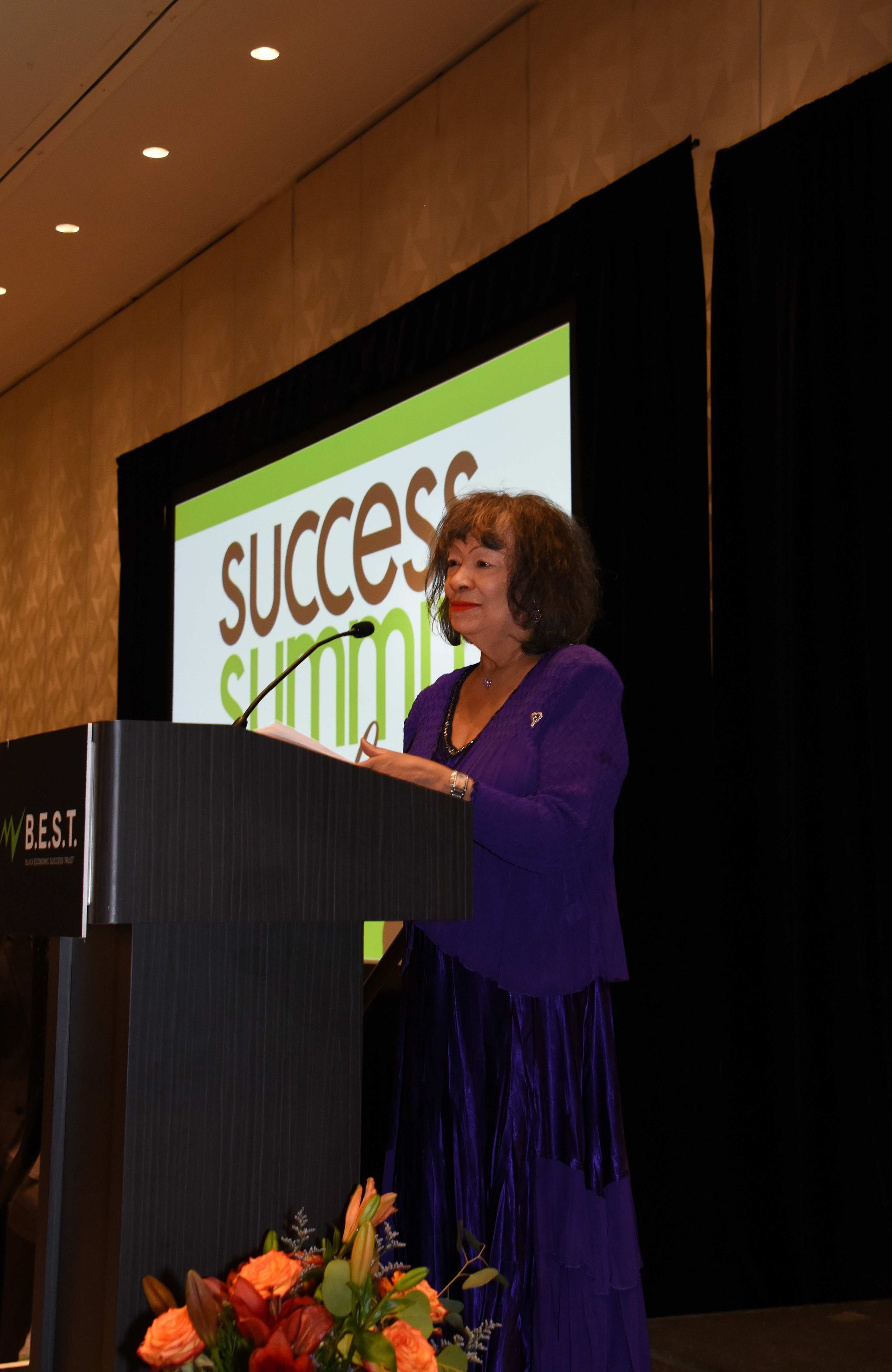 A woman in a purple dress stands at a podium in front of a screen that says success