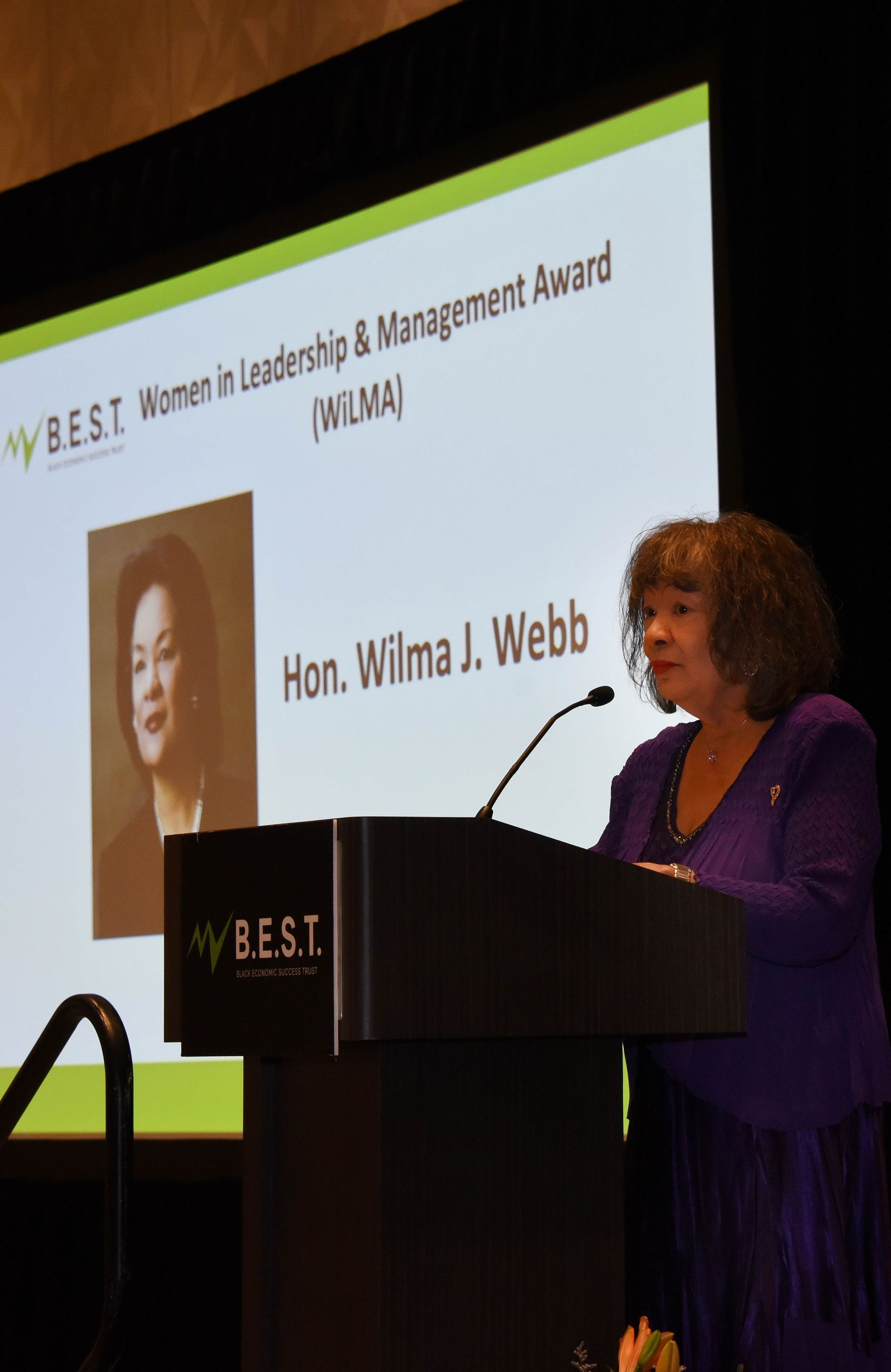 A woman stands at a podium in front of a screen that says hon wilma j webb