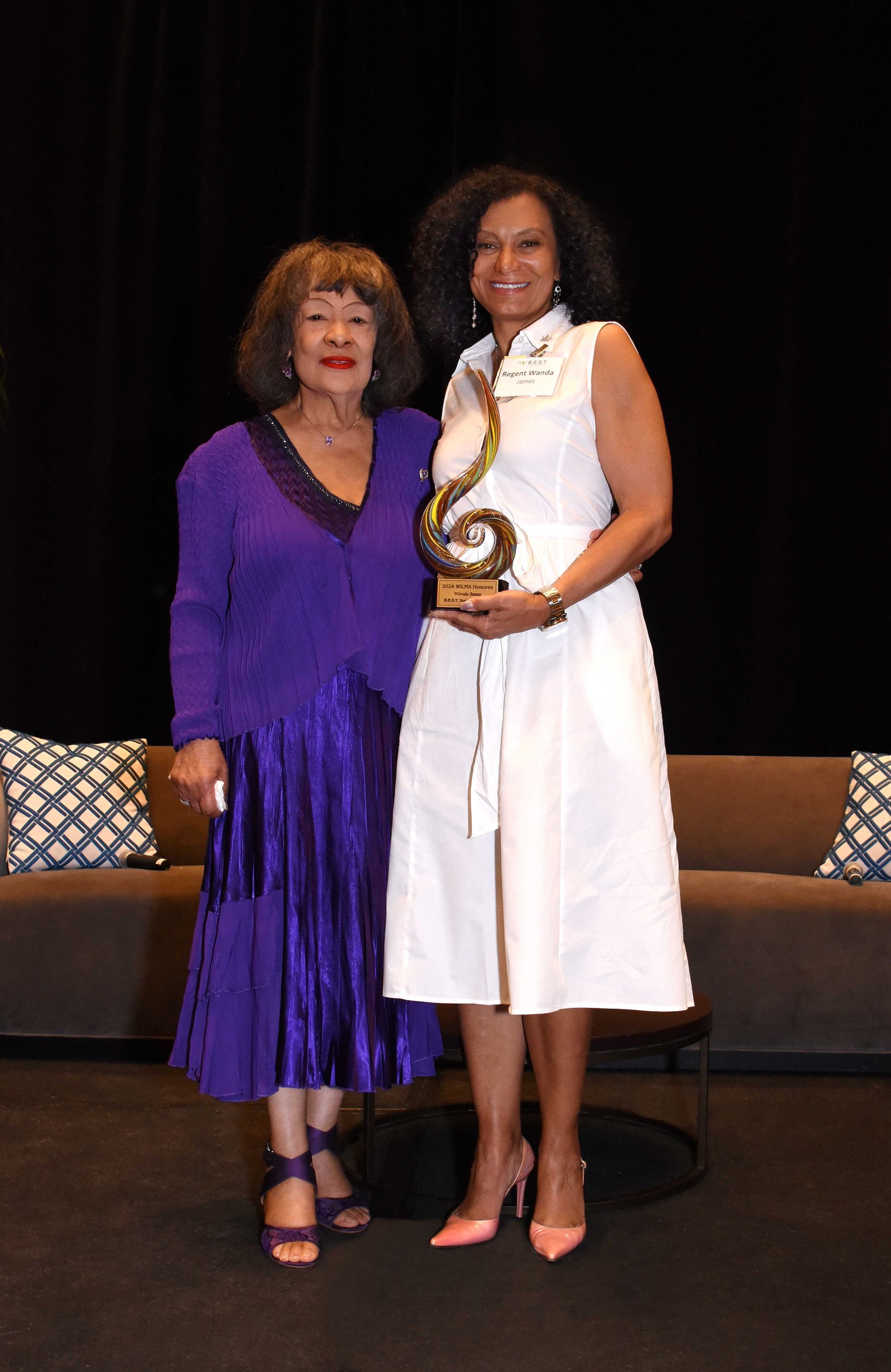 Two women are standing next to each other on a stage holding a trophy.