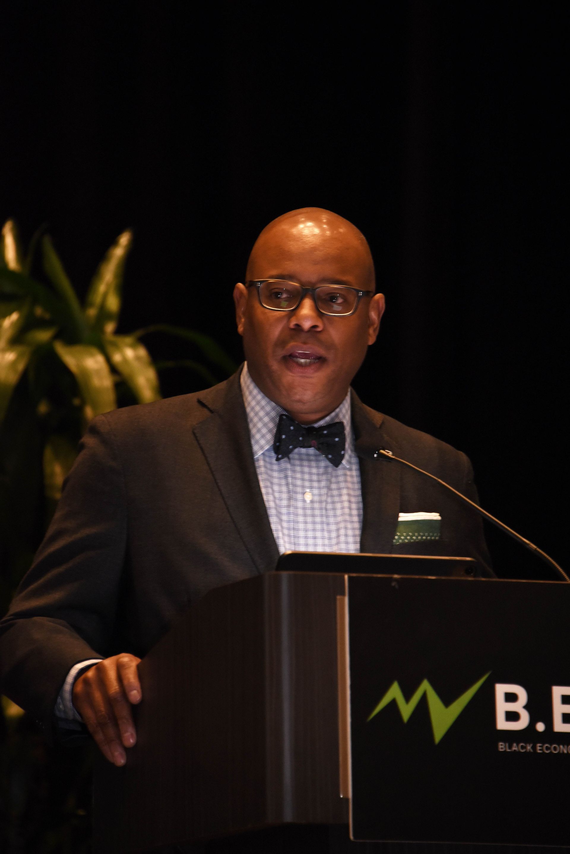 A man in a suit and bow tie is giving a speech at a podium that says b.e.