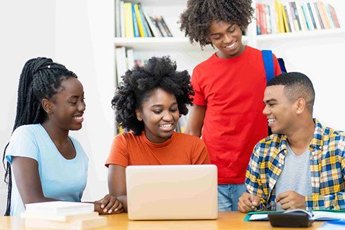 A group of young people are sitting around a table looking at a laptop computer.