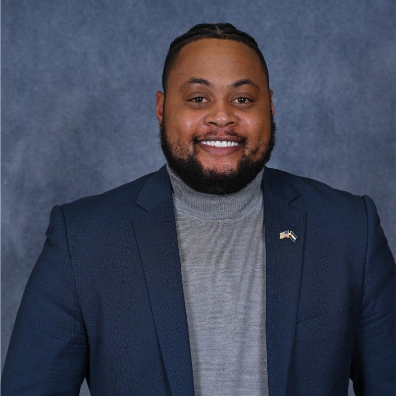 A man with a beard is wearing a tan suit and smiling.