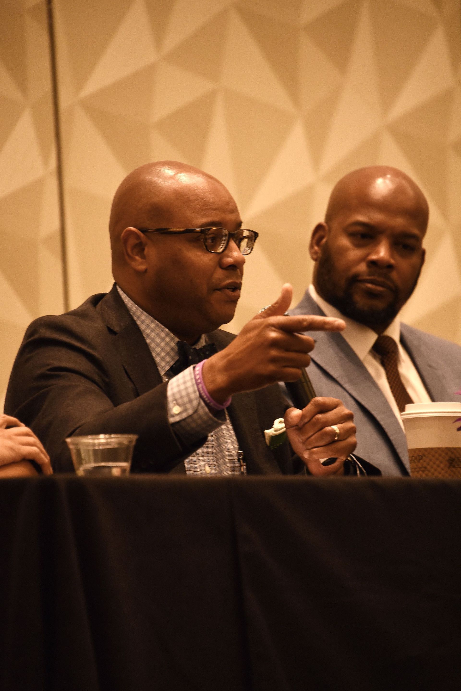 A man in a suit and tie is sitting at a table talking into a microphone.