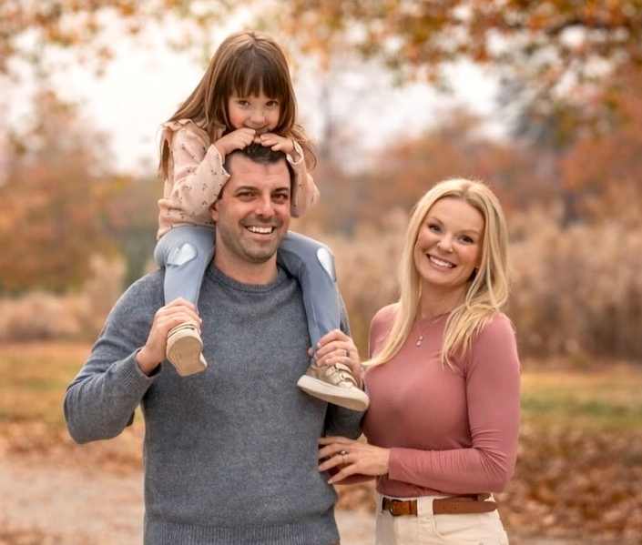 Family of three smiling outdoors: father with child on shoulders, mother beside them. Autumn foliage.