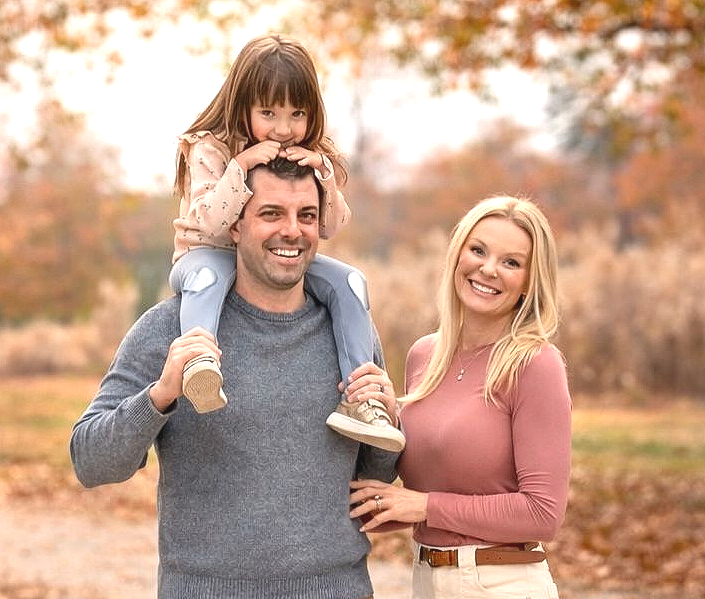 Family of three smiling outdoors: father with child on shoulders, mother beside them. Autumn foliage.