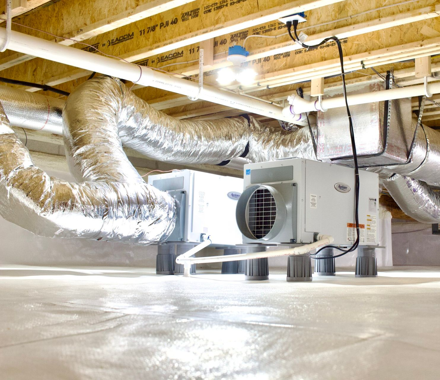 Crawl space with HVAC system, ductwork, and lighting under a home, sitting on a white moisture barrier.