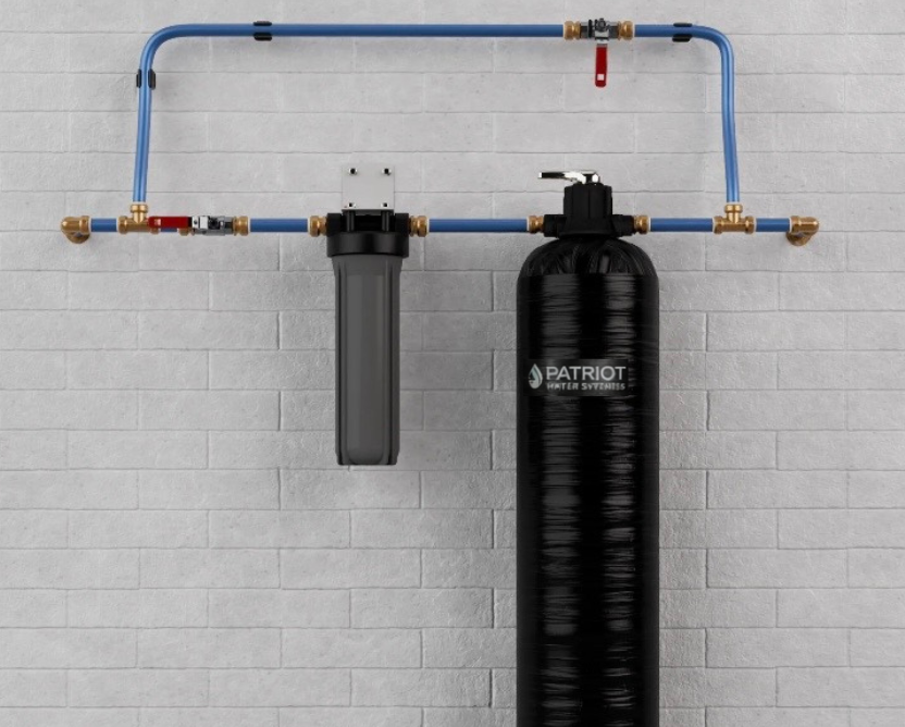 A water filtration system with a sediment filter and a large black tank connected by blue piping against a brick wall.