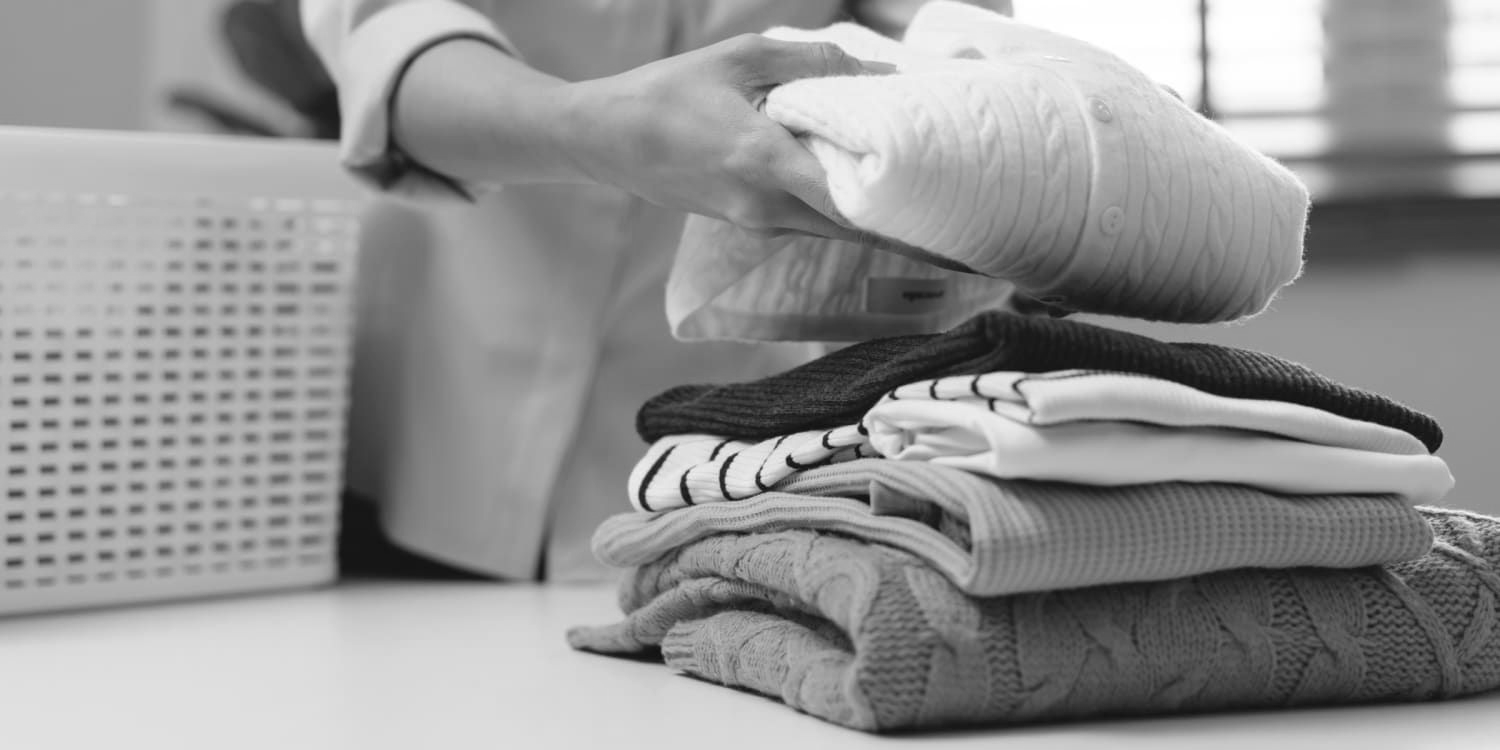 A person folding a stack of clean clothes on a table next to a laundry basket.
