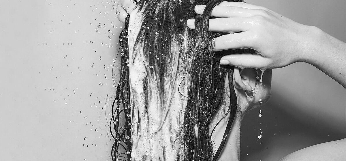 A person rinsing shampoo out of their hair under a shower, with water droplets visible.