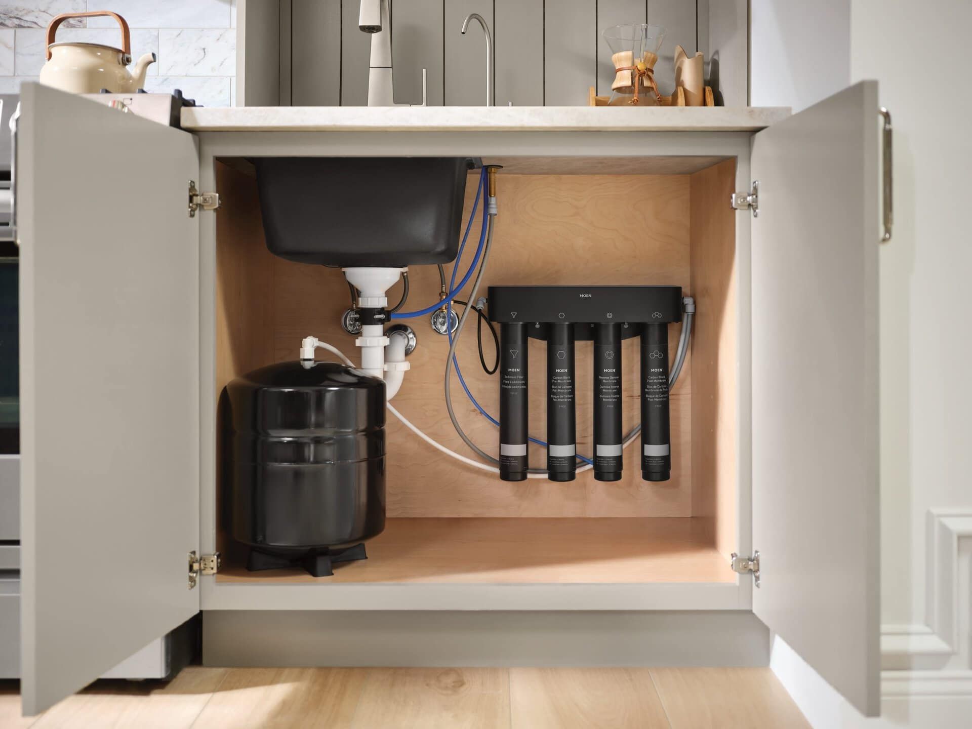 Under-sink water filtration system with a black tank and four connected cylindrical filters inside a kitchen cabinet.