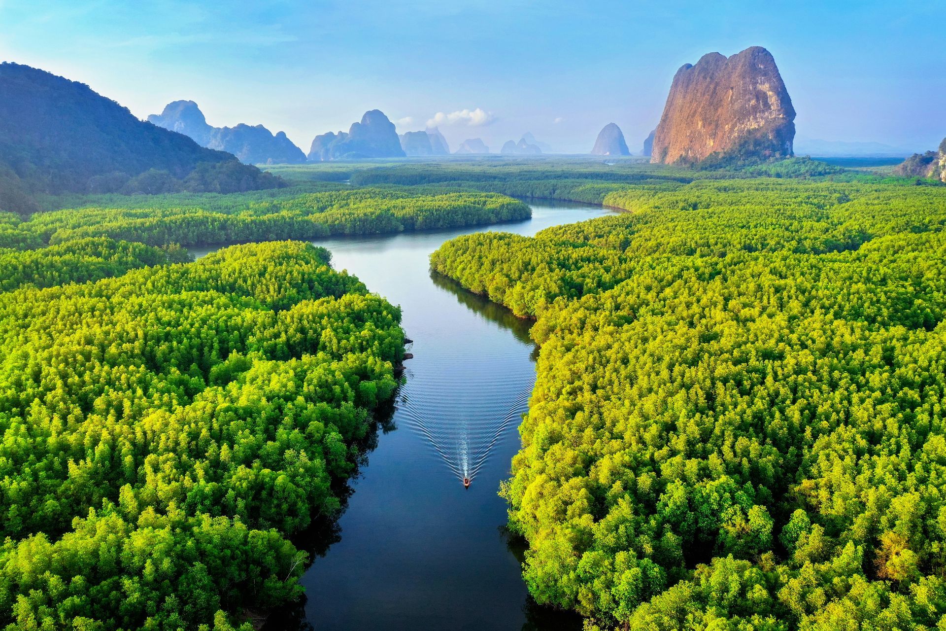 River winding through dense green mangrove forest, with limestone cliffs in the background under a blue sky.