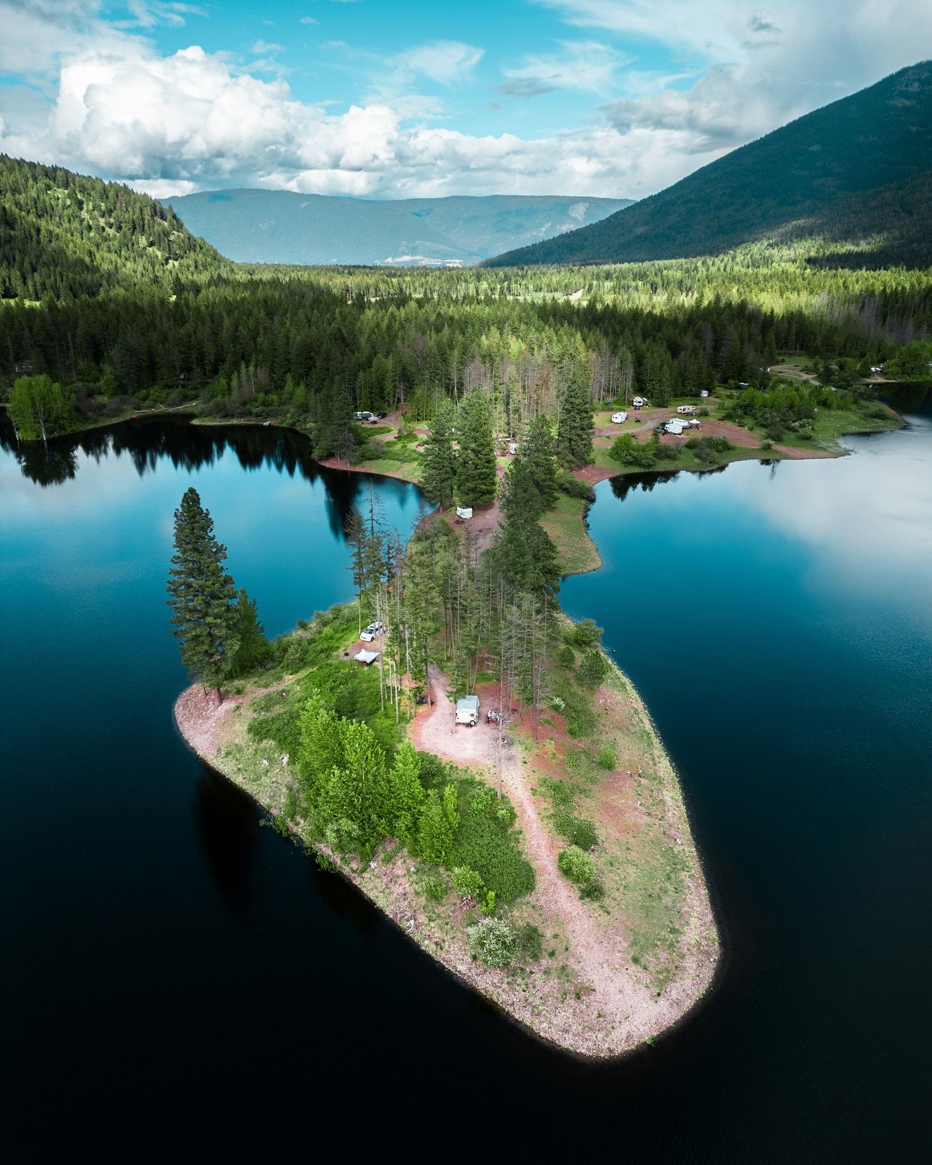 Island in a lake with trees and camping area. Mountains and green forest in the background. Blue water.