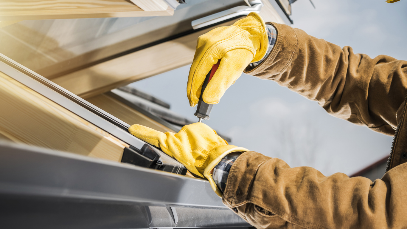 A person wearing yellow work gloves uses a screwdriver to install a window frame into a roof.