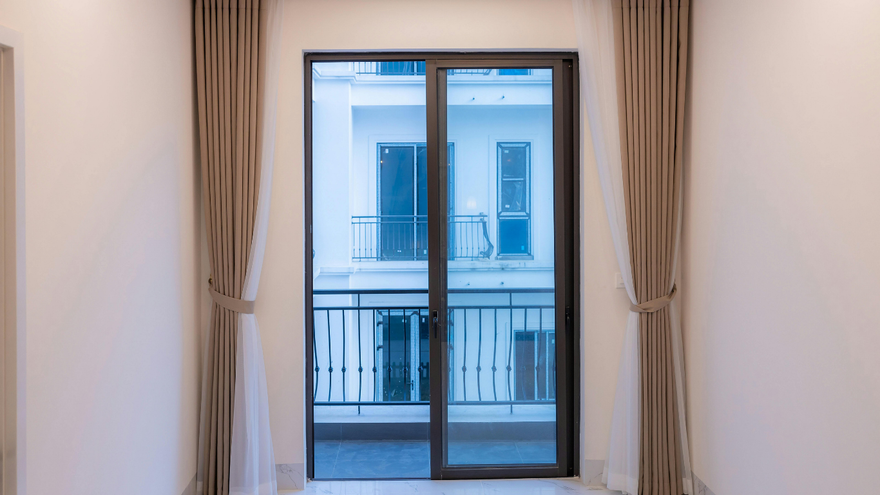 A balcony door framed by beige curtains, looking out toward a multi-story building across a street.