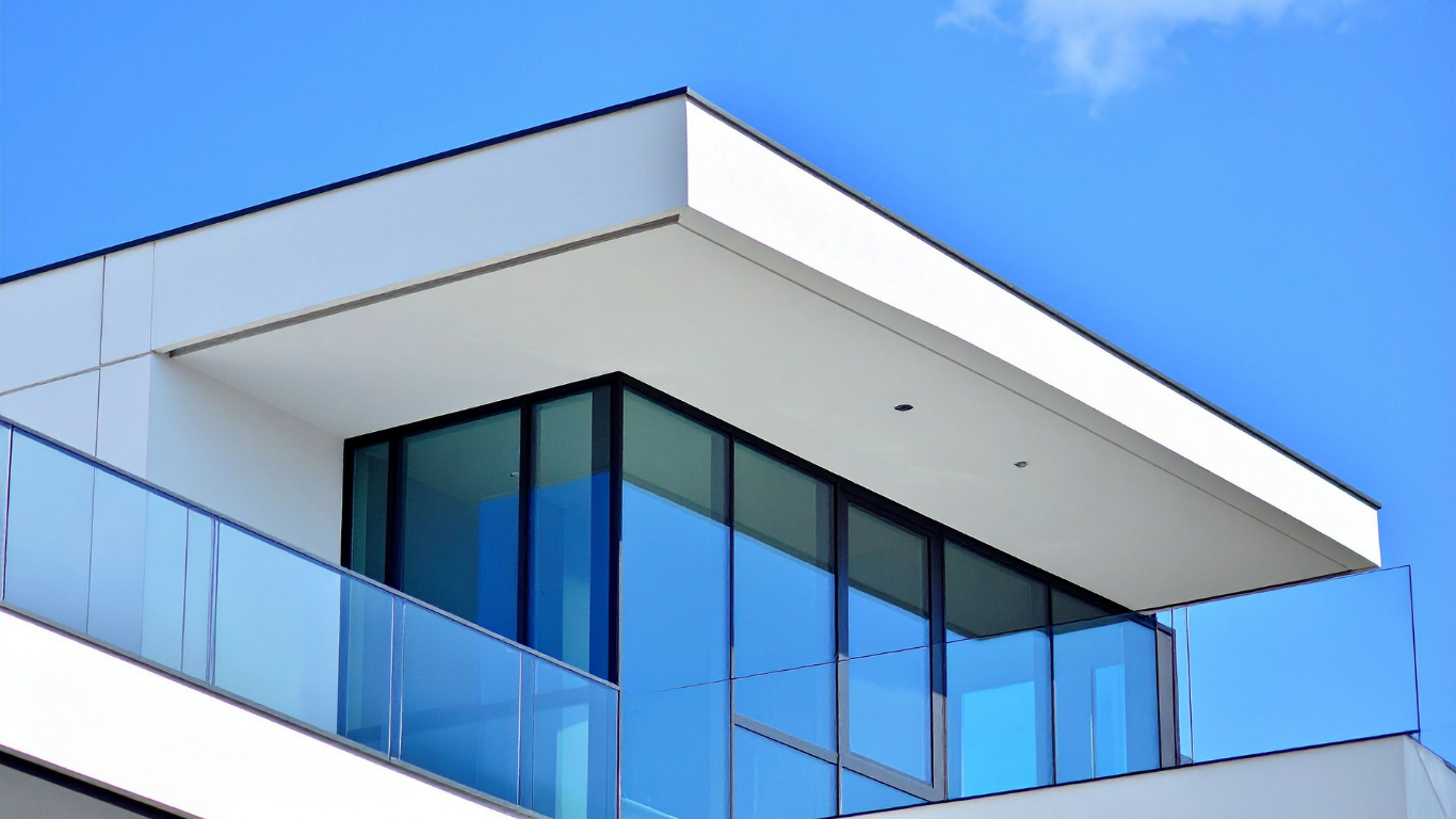 A glass-walled balcony and top-floor room with a white overhanging roof against a bright blue sky.