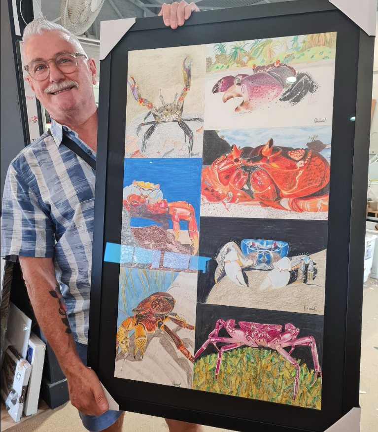 Dean is holding up a large painting of crabs with black frame around it — Next Level Picture Framing In Ballina, NSW