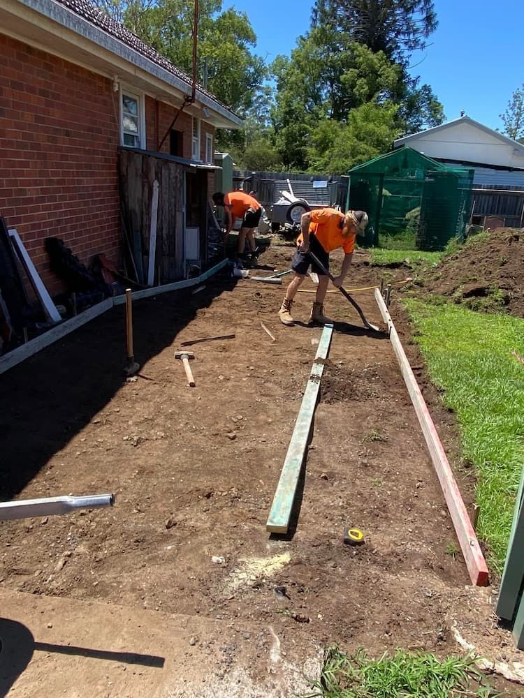 Two Workers in Orange Shirts Leveling Dirt in a Backyard, Alongside a Brick Building — JDC Concreting in Grafton, NSW