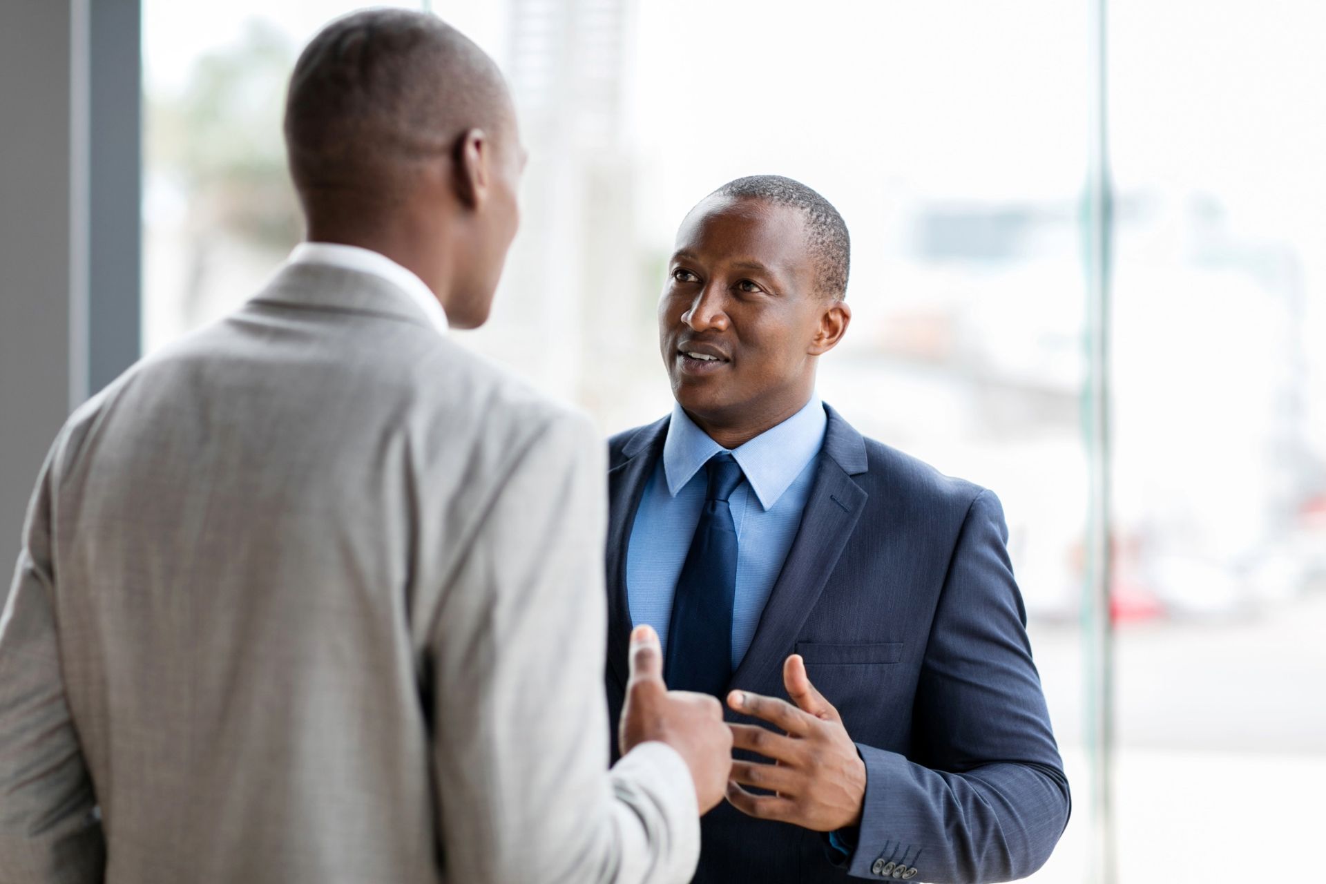 Two men in suits talking in an office setting. One points while speaking.
