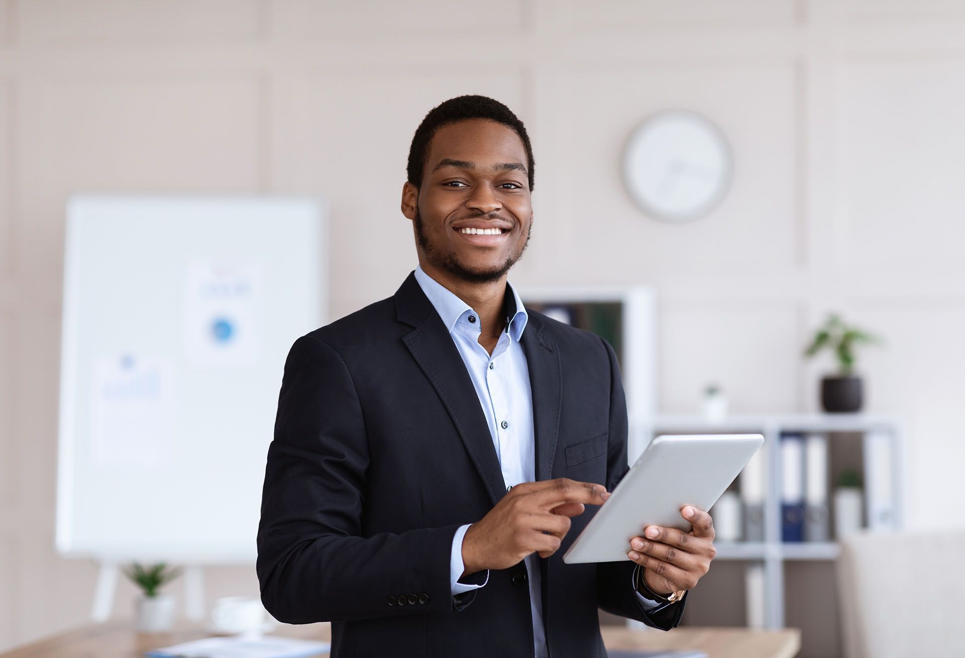 Smiling Black businessman in a suit holds a tablet in an office setting.