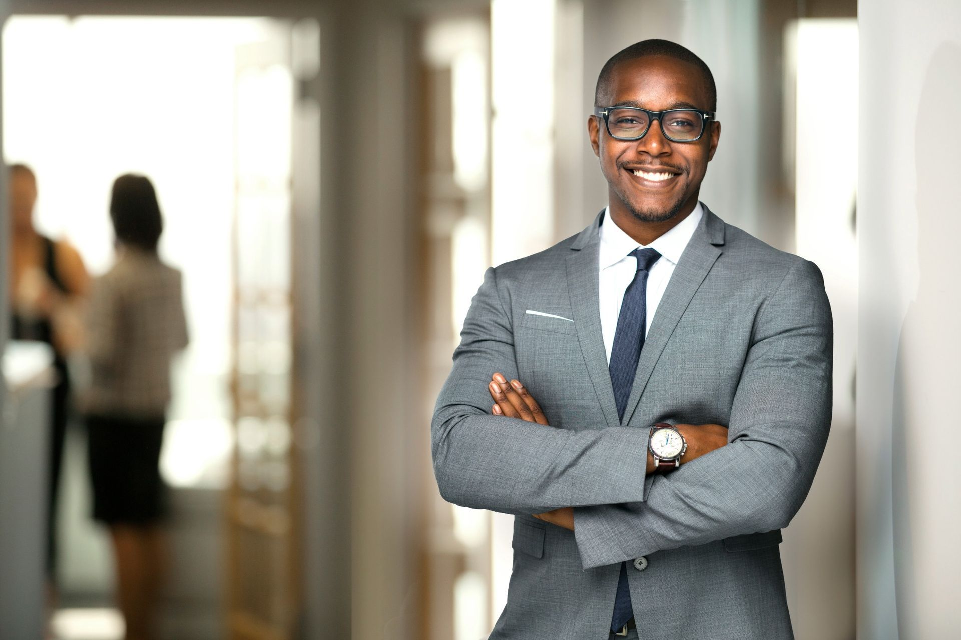 Man in gray suit and glasses, smiling with arms crossed in office hallway.