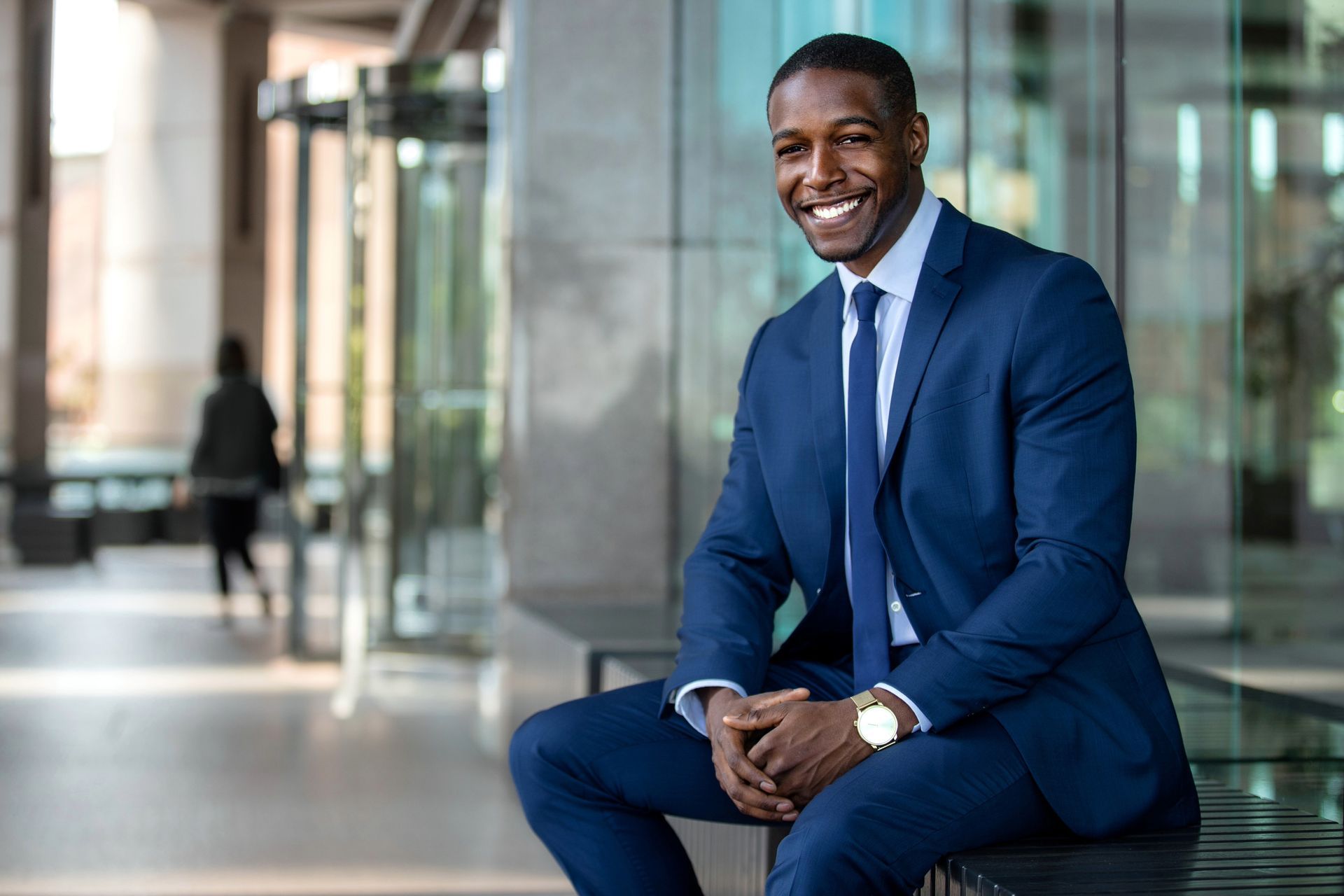 Man in a blue suit smiles, sitting outside a modern building.