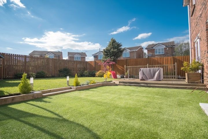A fenced-in backyard with a green lawn, tiered wooden borders, a small wooden deck, and neighboring houses under a blue sky.