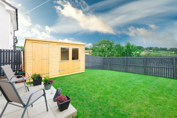 A backyard with a wooden shed, a green lawn, dark grey fences, and two patio chairs on a stone-tiled seating area.
