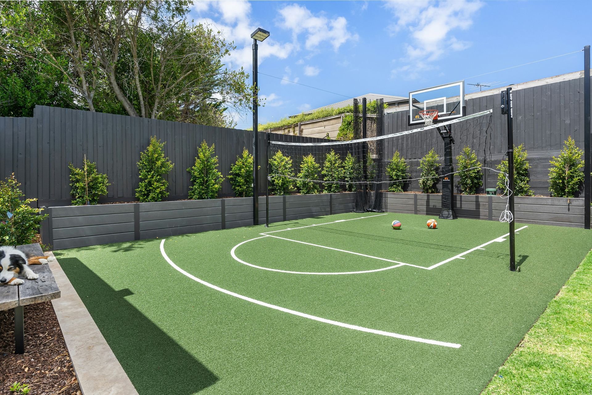 A backyard multi-sport court with artificial turf, basketball hoop, and volleyball net, surrounded by a dark grey fence.