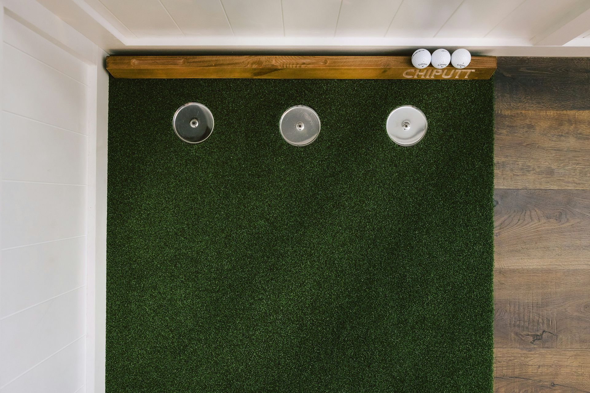 Top-down view of an indoor putting mat with three holes, a wooden frame, and three golf balls resting on the edge.