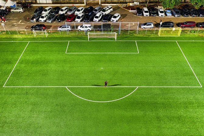 Aerial view of a soccer field with a lone person standing near the center line, adjacent to a parking lot.