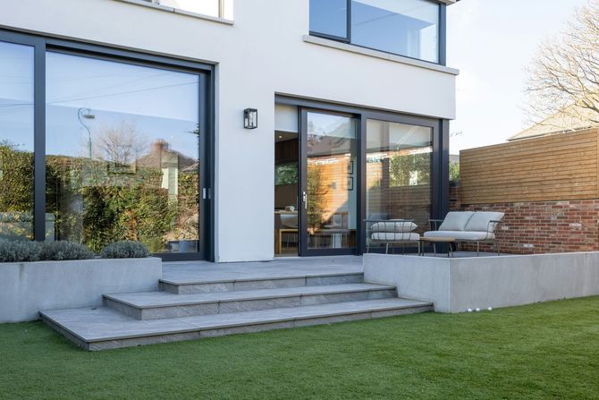 Modern white house with floor-to-ceiling glass doors, stone patio steps, and a minimalist outdoor lounge area.