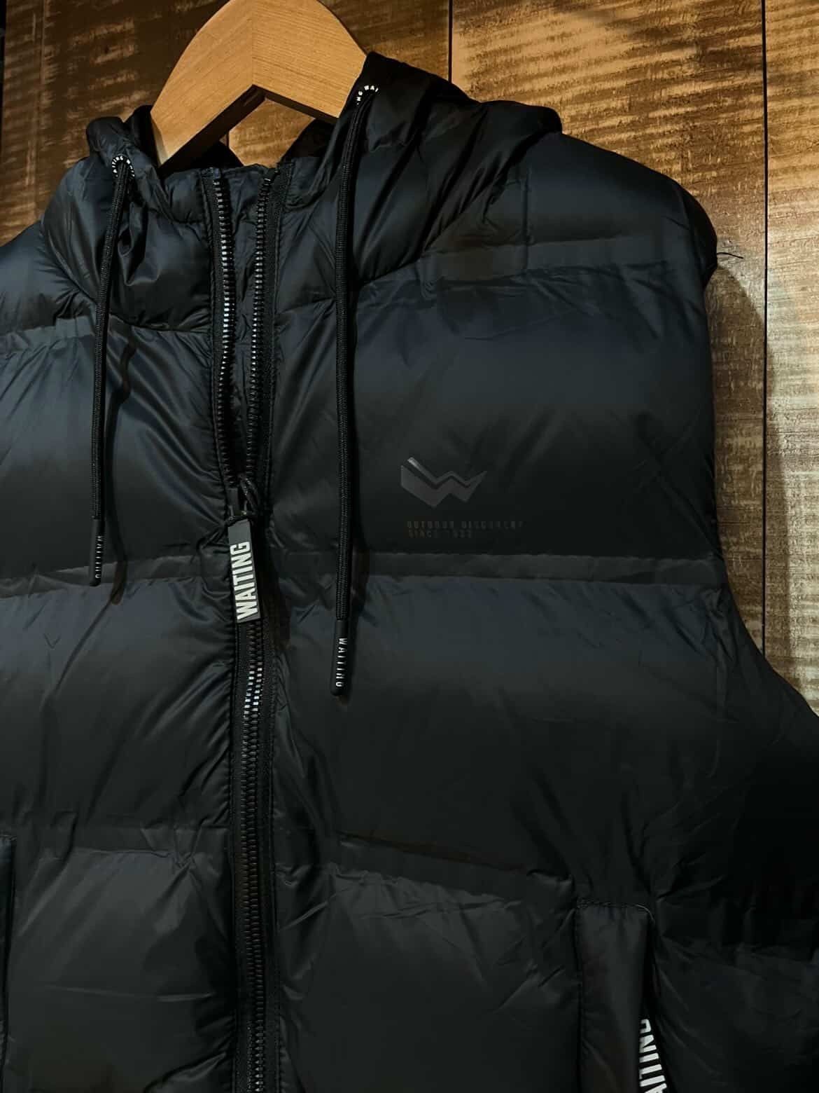 Chaleco acolchado negro sin mangas colgado de una percha de madera sobre un fondo marrón texturizado.
