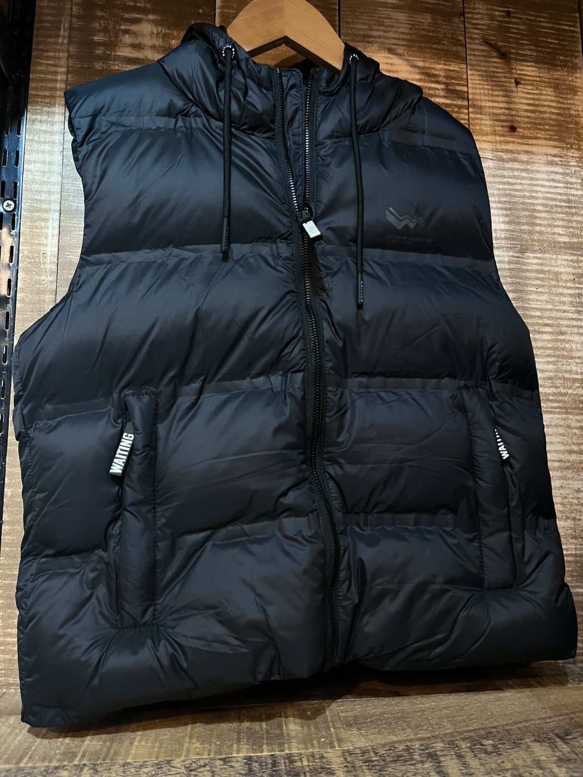 Chaleco acolchado negro sin mangas colgado de una percha de madera sobre un fondo marrón texturizado.