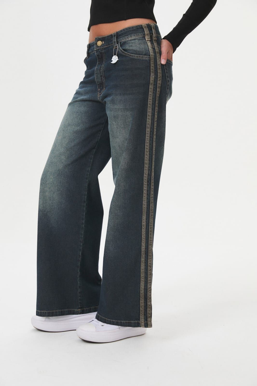 Persona con pantalones vaqueros anchos oscuros con rayas laterales blancas y zapatillas blancas sobre un fondo liso.