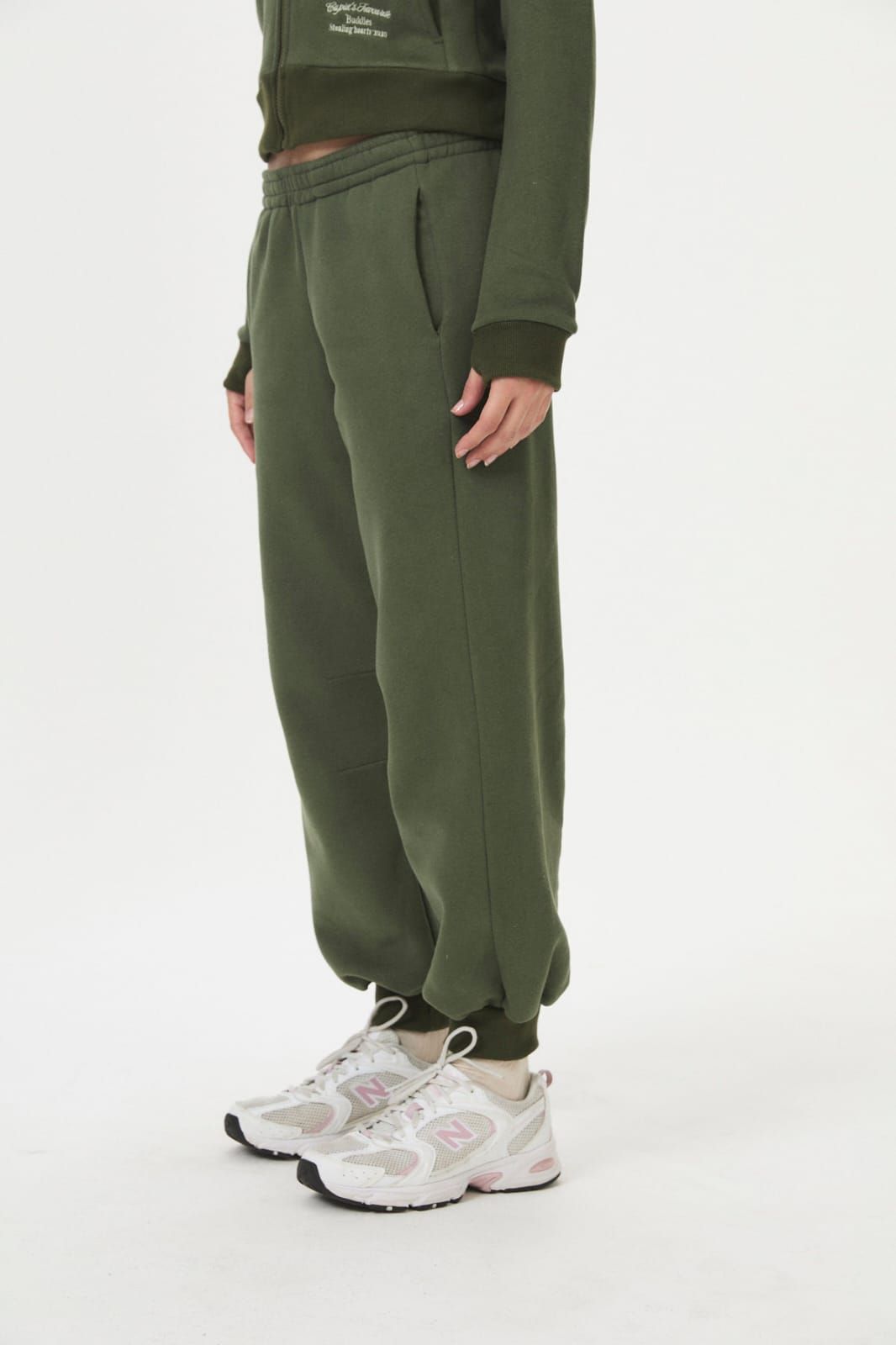 Persona con sudadera y pantalones deportivos de color verde oliva, con zapatillas blancas, de pie sobre un fondo blanco liso.