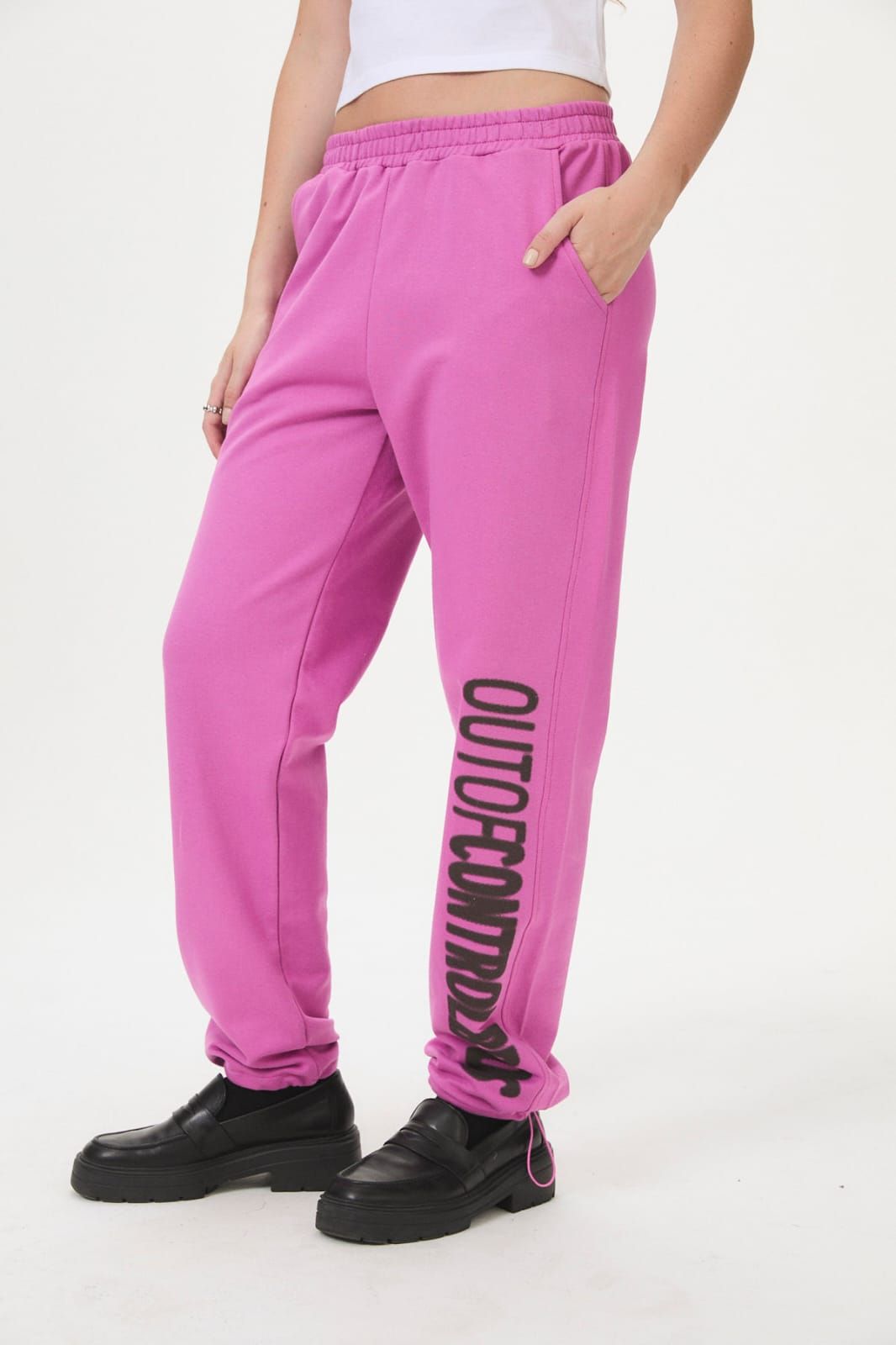 Persona con pantalones deportivos de color rosa brillante y zapatos negros, de pie sobre un fondo blanco.